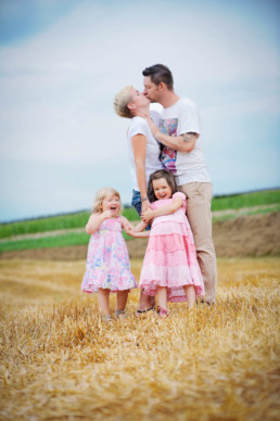 Familienfoto auf dem Feld, Eltern küssen, Kinder lachen